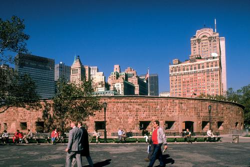 People crossing an open area in Battery Park, with the red sandstone ring of Castle Clinton and towering buildings set against a blue sky behind it.