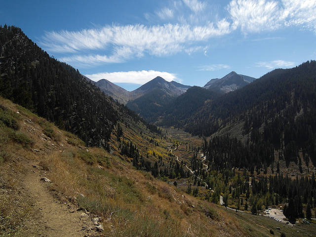 Multiple mountain peaks lie beneath the blue sky on a partly cloudy day, with trees and a narrow path sitting in the valley below.