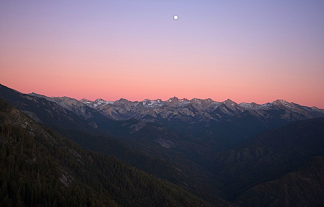 A long string of mountain peaks dig into the purple and orange sky at sunset. Toward the top of the image is a small full moon.