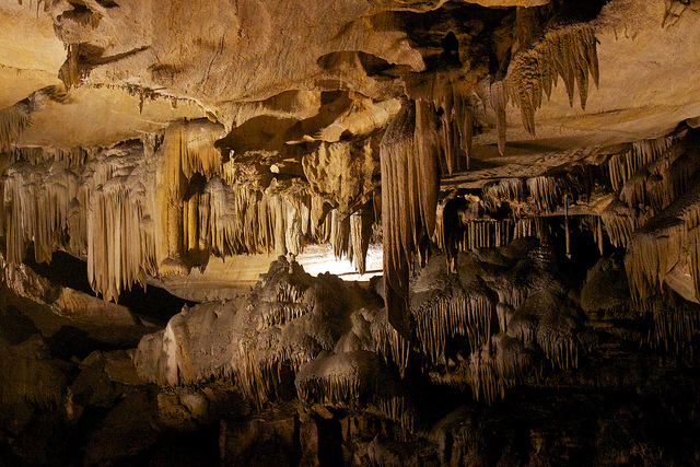 Innumerable stalactites hang down in clumps from inside the cave. The rock is primarily of a light brown hue.