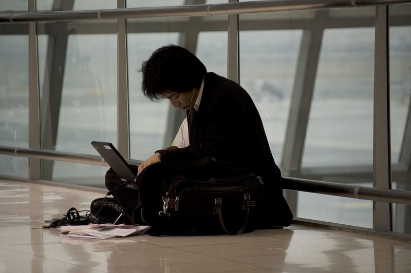 A man sits on the floor of an airport, using his laptop. 
