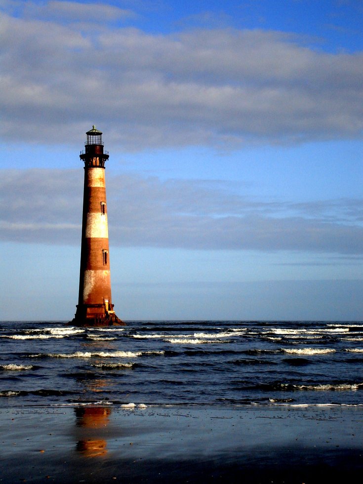 Photo taken from beach of faded brown and white faded lighthouse cast with hues of afternoon light sitting out at sea