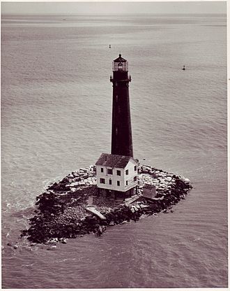 Pictured is a tall, slim black lighthouse with a granite base in the middle of the Gulf Coast on a small rocky island. 