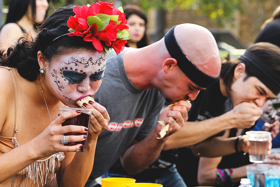 Woman with a skull painted on her face competes in Chacho's taco eating contest next to other competitors including Matthew Stonie. 