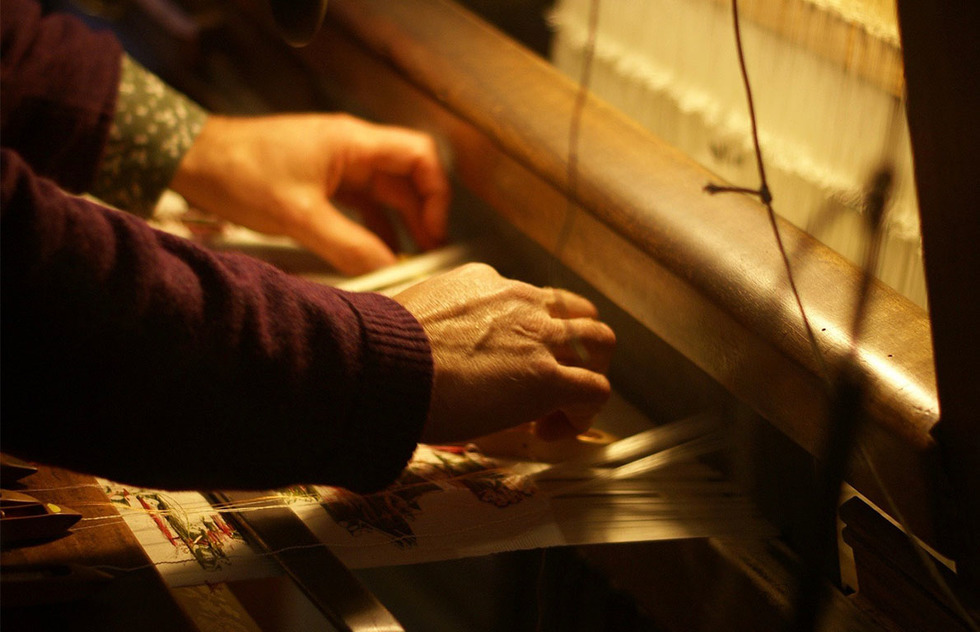 A silk worker using both hands to weave a patterened textile on a large Jacquard loom