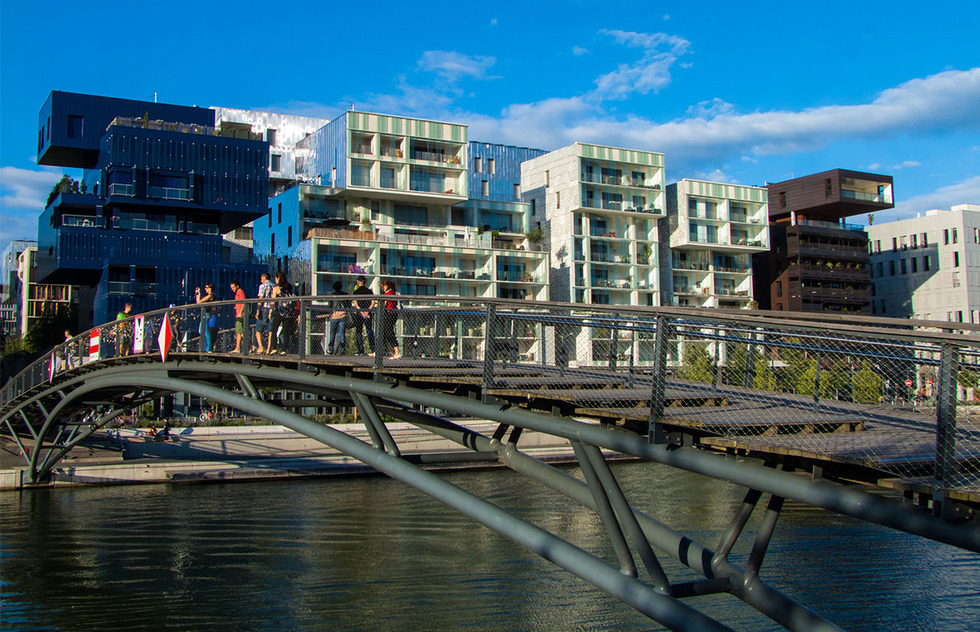 Pedestrians crossing over a skinny bridge at the Confluence to the colorful cubic apartment buildings covered in large windows