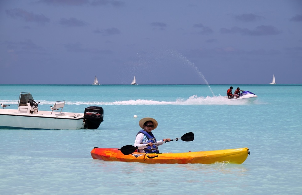A woman kayaking in open water at Jolly Beach, Antigua. Behind her are an empty speedboat, two boys on a jet ski, and three sailboats in the distance.
