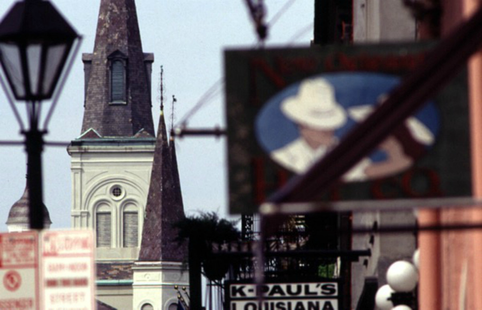 A raised level street view captures the old-fashion intricacy of the New Orlean's French Quarter. Behind the old black lamp post and hanging restaurant sign, lies the towering steeple of an old white church. 
