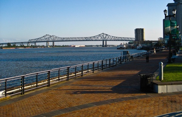 A smooth brown promenade winds down along the rolling Mississippi, forming the Moon Walk of Woldenberg Park. The view includes the buildings along the New Orleans river and a distant bridge.  