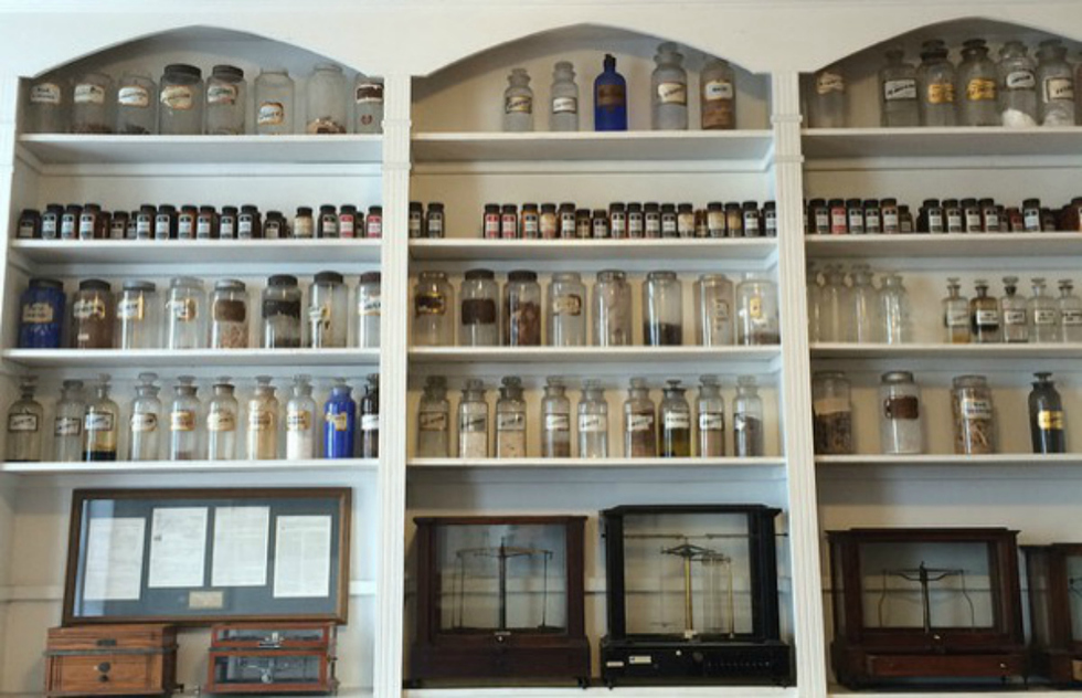 This photo features a series of shelves filled with old-fashioned, glass medical bottles. Below are well-preserved balance scales in their cases. 