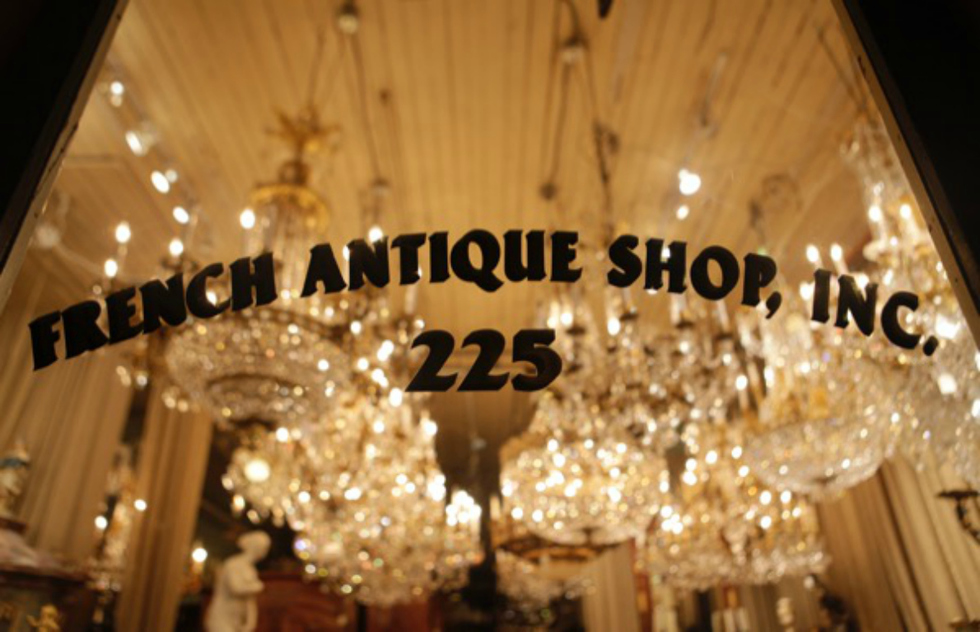 A clean glass window store front is illuminated by the dazzling chandeliers, showing the etched black lettering—French Antique Shop. 